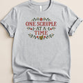 A gray short-sleeve t-shirt features cross-stitch style text that reads ONE SCRUPLE AT A TIME, surrounded by floral and mortar graphics.