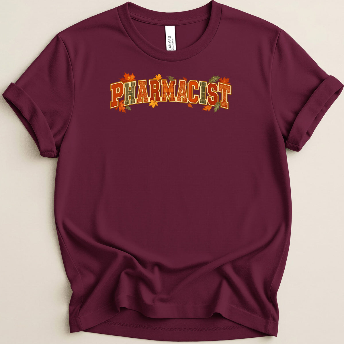 A maroon t-shirt featuring the word Pharmacist in autumn-colored block letters with autumn leaves.