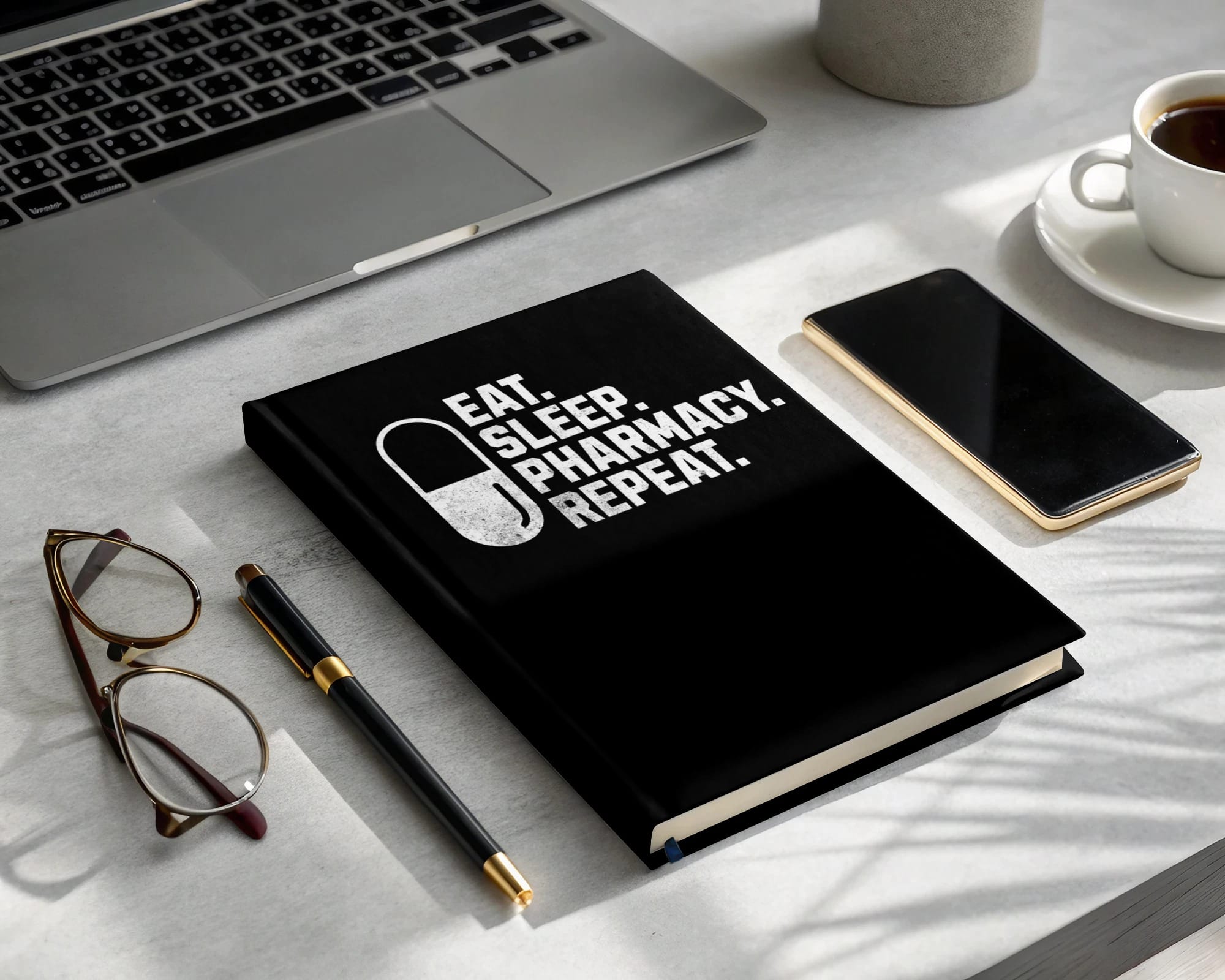 Black notebook with "EAT. SLEEP. PHARMACY. REPEAT." text, laptop, glasses, pen, and coffee.
