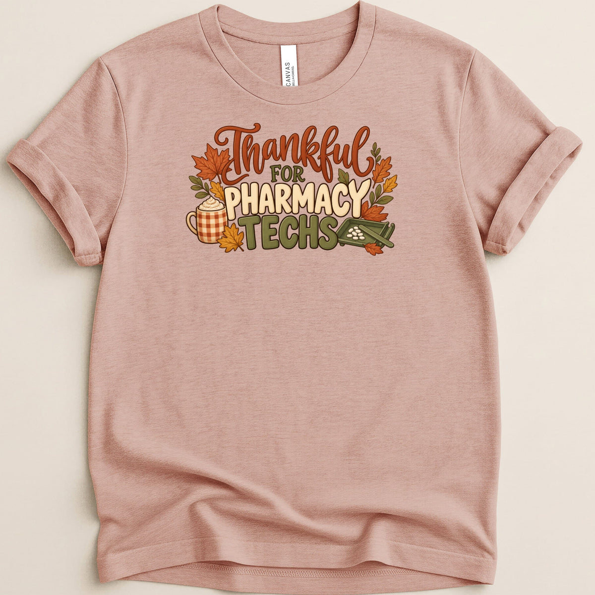 A dusty pink crewneck t-shirt with rolled sleeves featuring the text "Thankful for Pharmacy Techs" surrounded by autumn leaves, a coffee mug, and a pill counting tray.