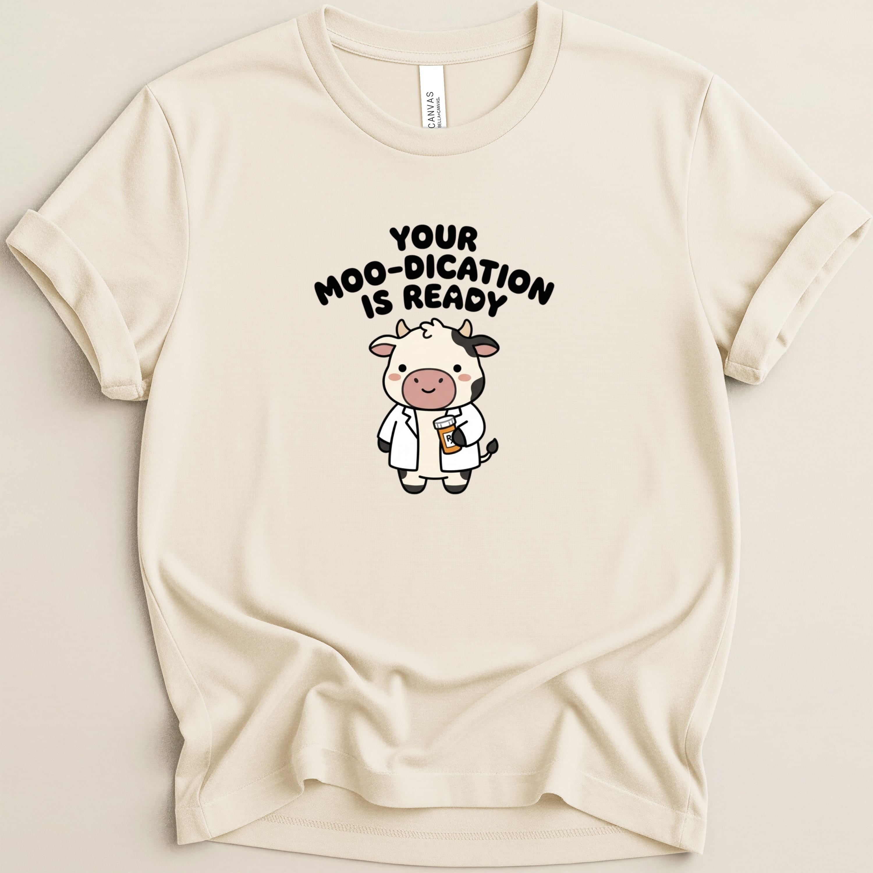 Cream t-shirt with cartoon cow wearing a lab coat, holding a pill bottle, text reads "Your Moo-dication Is Ready".
