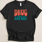 Black t-shirt with "DRUG EXPERT" text in distressed orange and teal lettering.