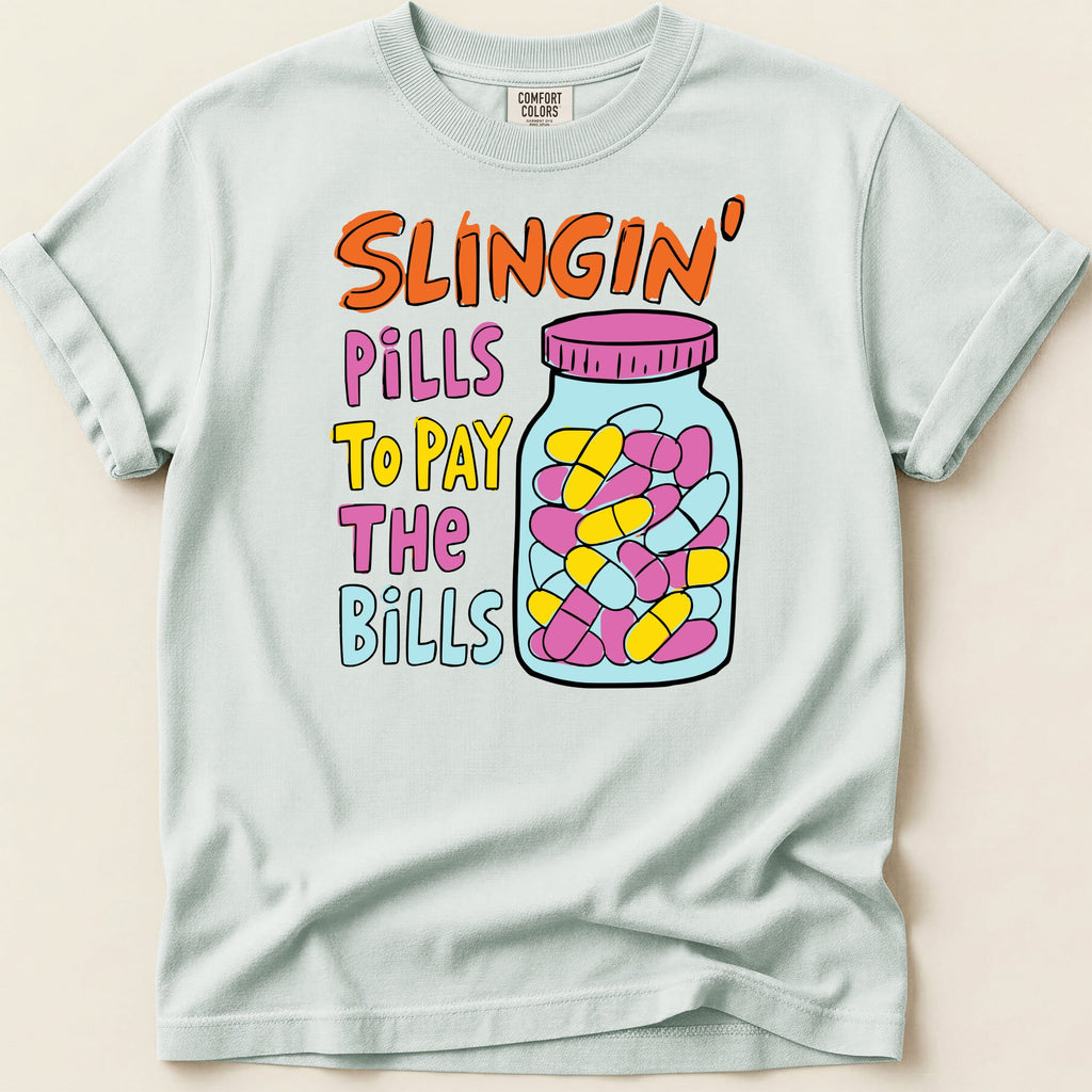 Light blue Comfort Colors t-shirt with "Slingin' Pills To Pay The Bills" text and jar of pills graphic.