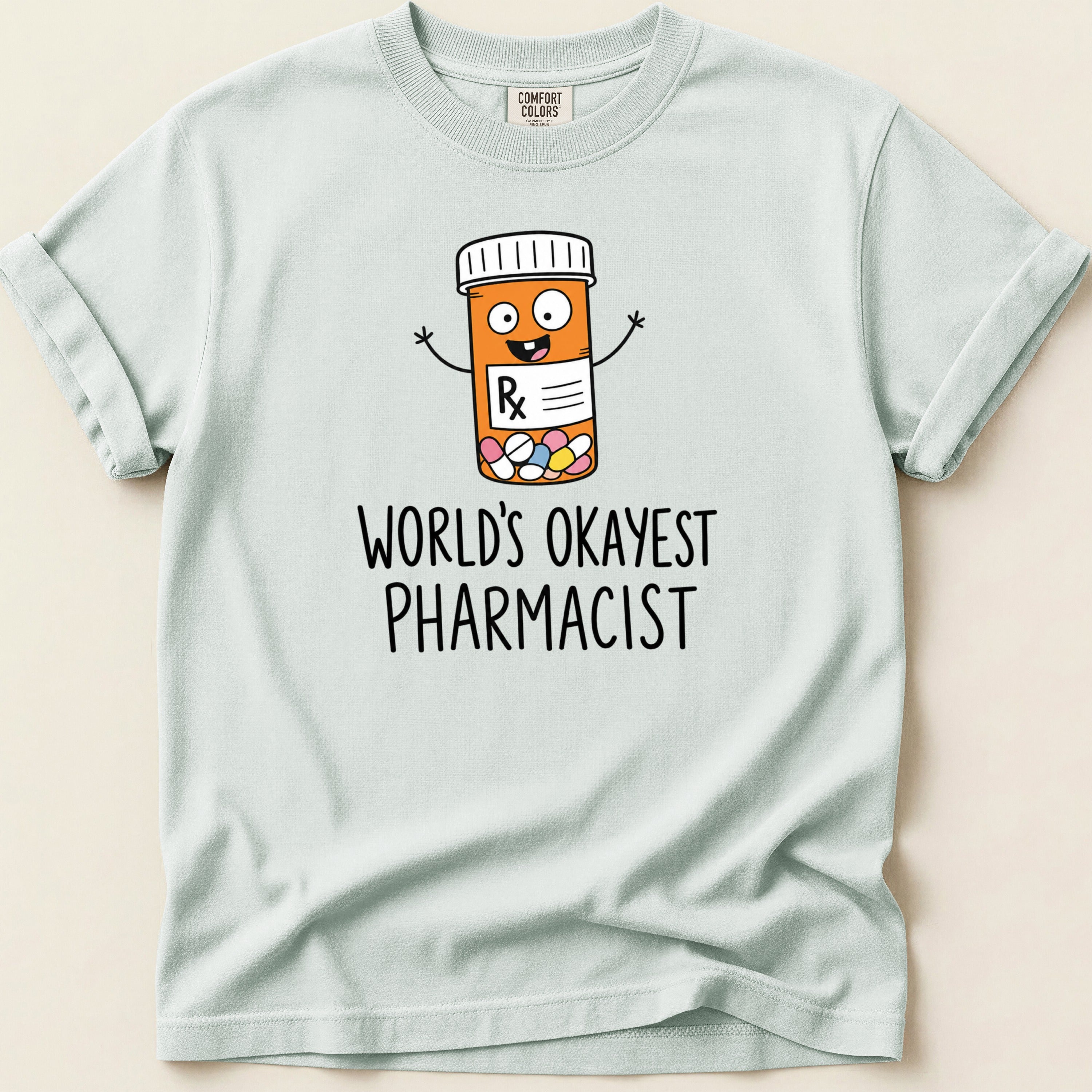 Light blue Comfort Colors shirt with a cartoon pill bottle and "World's Okayest Pharmacist" text.