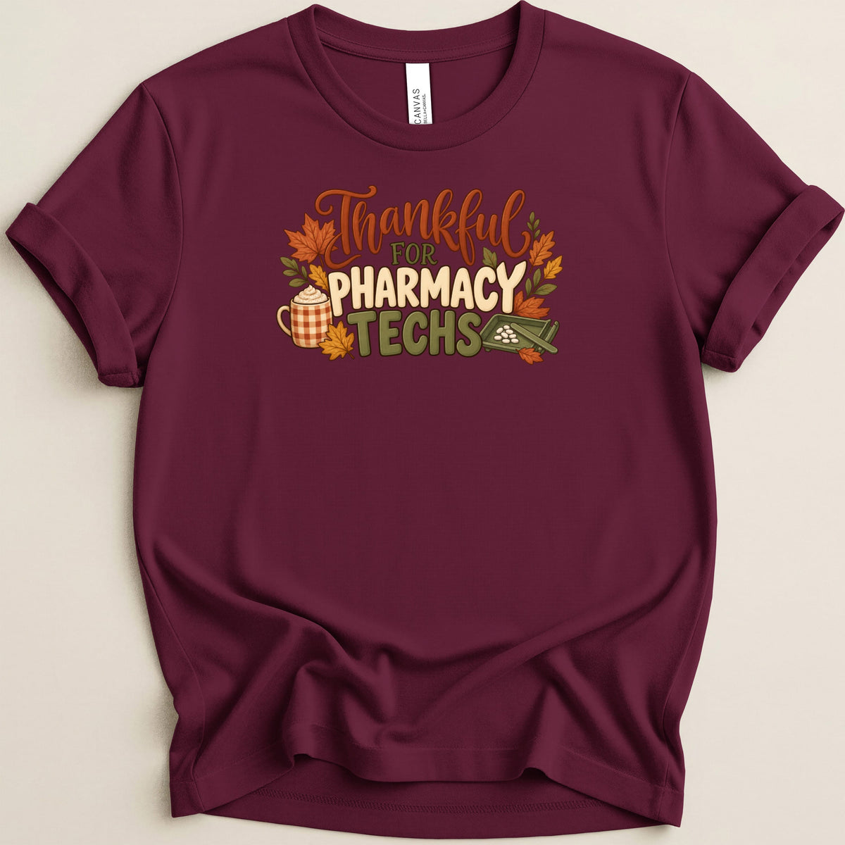 A maroon crewneck t-shirt featuring the autumn-themed text "Thankful for Pharmacy Techs" surrounded by fall leaves, a coffee mug, and a pill counting tray.