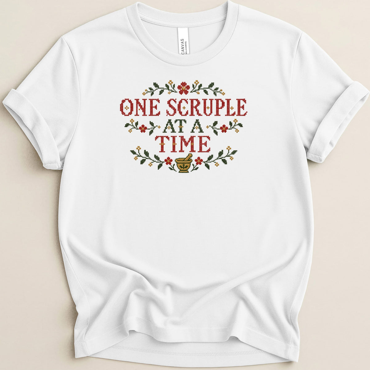 White short-sleeved t-shirt with red and green cross-stitch design reading One Scruple At A Time above a mortar and pestle.