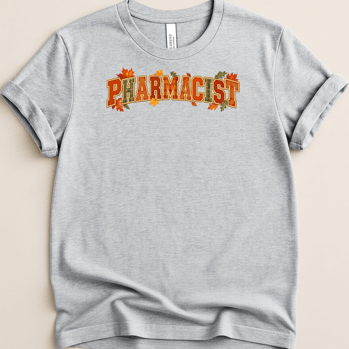 A light gray crewneck t-shirt with rolled sleeves featuring the word Pharmacist in orange block letters decorated with autumn leaves.