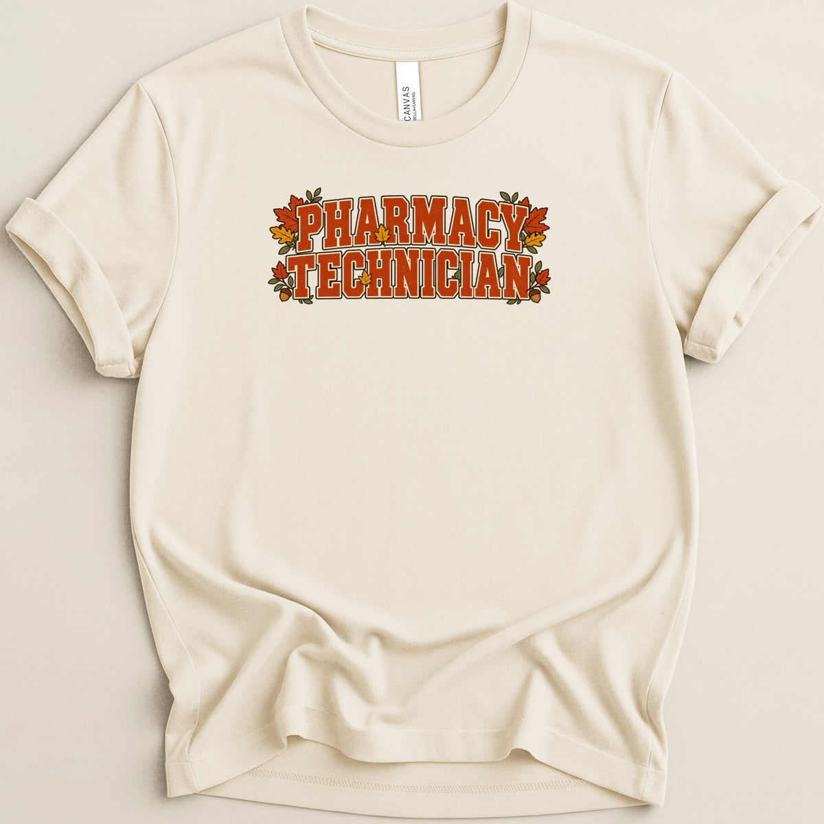 A beige crewneck t-shirt with rolled sleeves features orange autumn-themed text reading Pharmacy Technician.