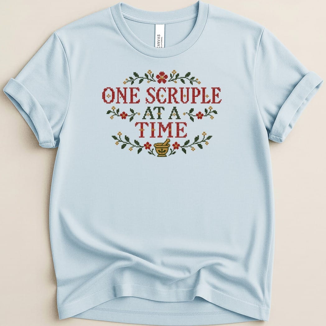 Light blue crewneck t-shirt featuring cross-stitch embroidery reading ONE SCRUPLE AT A TIME with floral motifs.