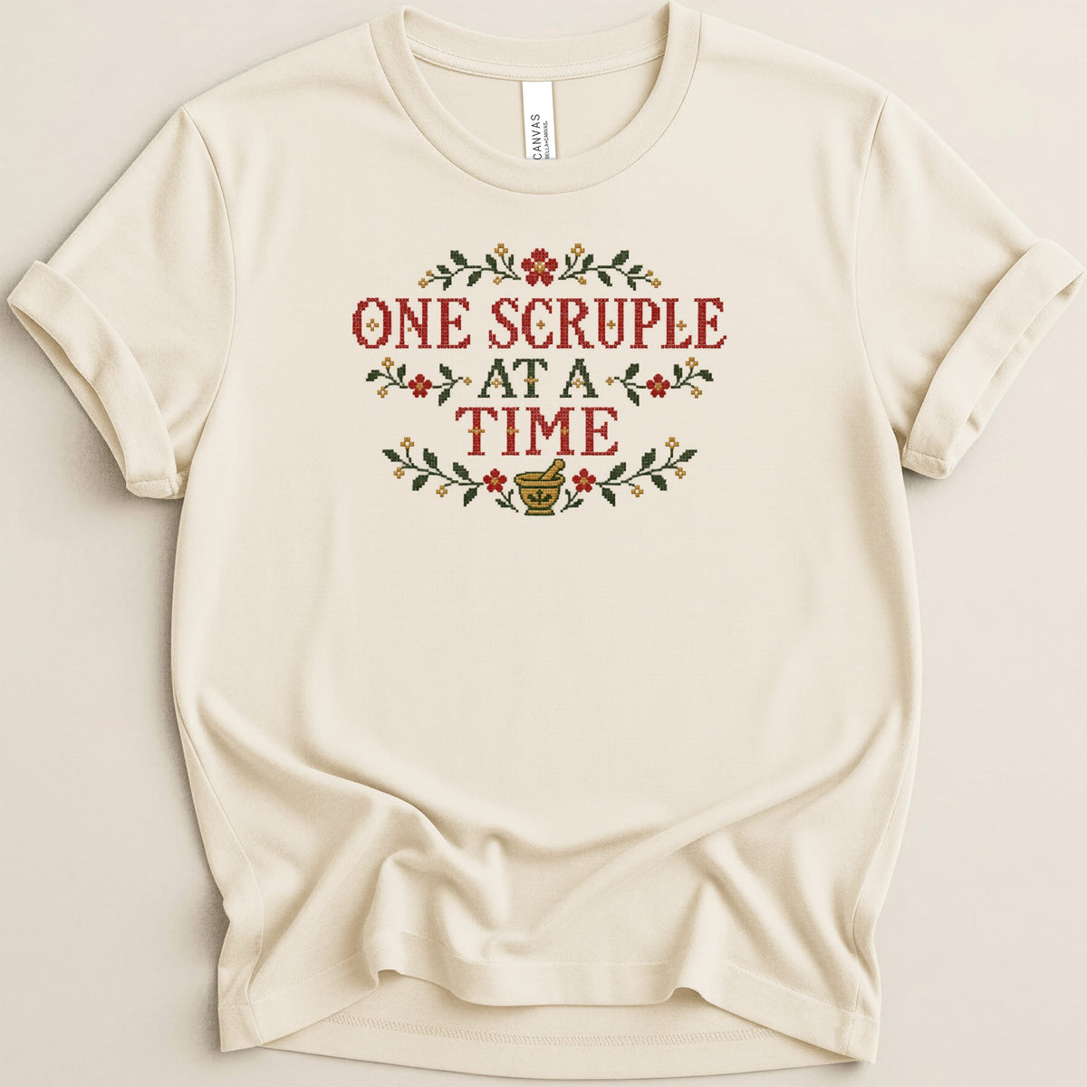 Cream crewneck t-shirt featuring cross-stitch embroidery that reads One Scruple At A Time surrounded by floral and mortar motifs.