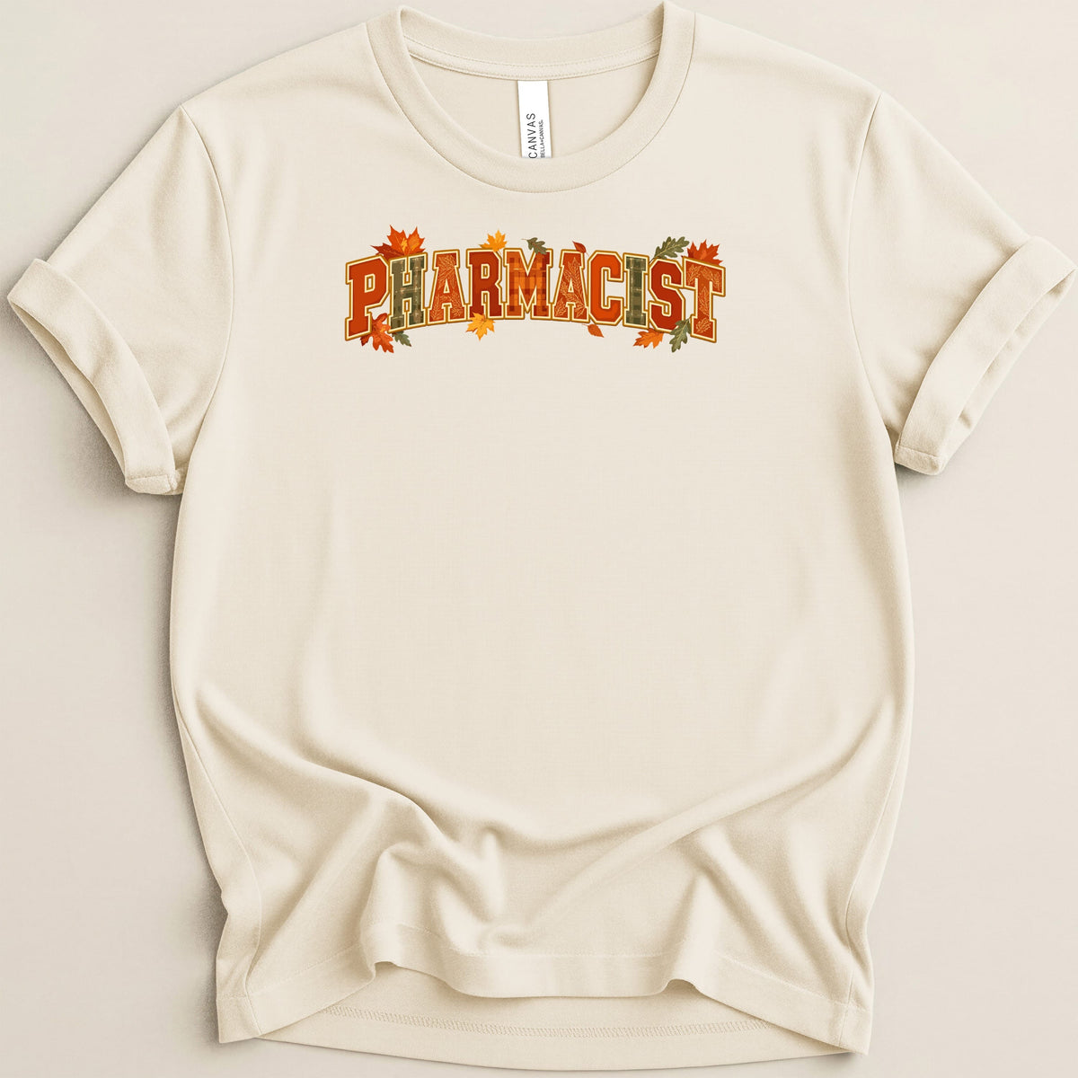 Cream-colored short-sleeve t-shirt with rolled cuffs, featuring the word PHARMACIST in orange block letters decorated with fall leaves.